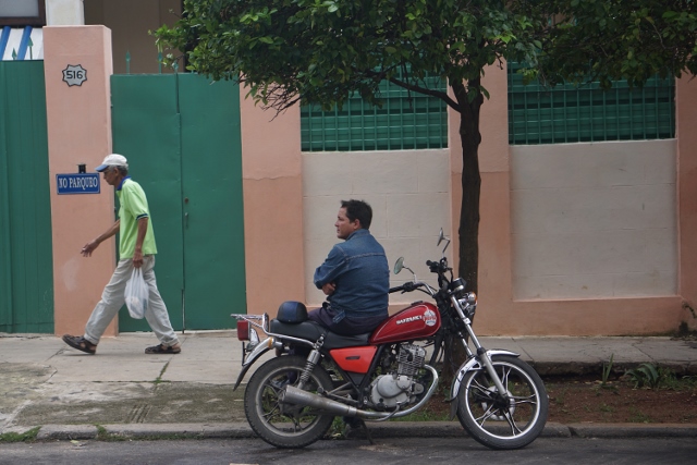 Man sitting on a parked motorcycle watching another man walk by.
